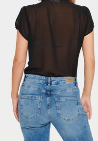 Blusa nera trasparente con maniche corte, abbinata a jeans denim azzurri con etichetta color cuoio in vita e due tasche posteriori.