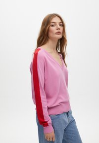 Rosa V-Ausschnitt-Pullover mit roten Seitestreifen. Weiches Material, gerippte Bündchen und Saum. Kombiniert mit hellblauen Jeans mit Perlenakzenten.