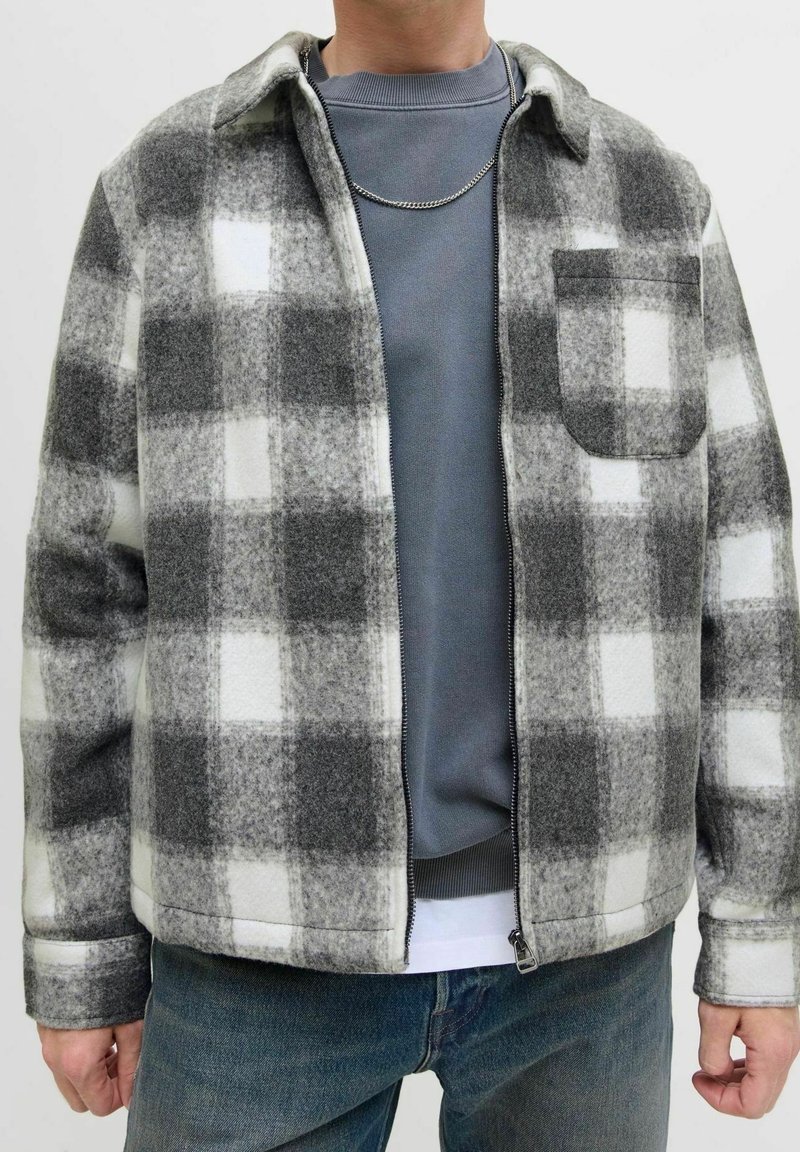 Personne portant une veste zippée à carreaux gris et blancs sur un sweat-shirt bleu et un jean bleu, avec un collier chaîne en argent visible.