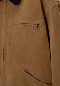 Veste en toile marron avec un col noir, une poche poitrine zippée horizontale avec une tirette perlée, et une poche inférieure avant inclinée.