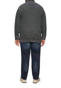 Zwarte denimjack met een gestructureerde afwerking, voorzien van een hoge kraag en geribde manchetten, gecombineerd met een donkerblauwe jeans en lichtgekleurde schoenen.