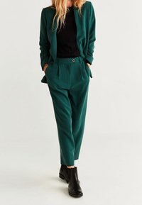 Completo su misura verde con giacca aderente e pantaloni affusolati. Il tessuto ha una consistenza liscia e presenta dettagli con bottoni dorati. Indossato con stivali neri.