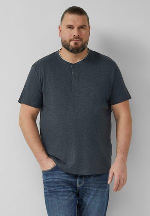 Mann mit Bart und zurückgebundenen Haaren, der ein dunkelgraues Henley-Shirt mit kurzen Ärmeln und blaue Jeans trägt, steht vor einem schlichten hellen Hintergrund.