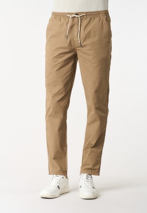 Pantaloni casual marrone chiaro da uomo con vita elastica e coulisse bianca, abbinati a sneakers bianche su sfondo semplice.
