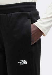 Χέρι στην τσέπη μαύρου παντελονιού North Face με ελαστική μέση, φορώντας άσπρο μακρυμάνικο πουκάμισο σε απλό φόντο.