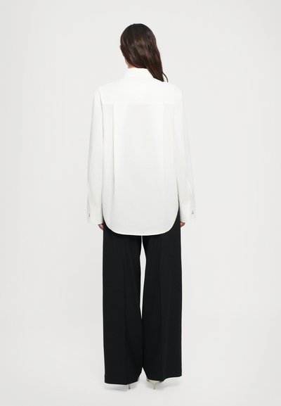 Camicia bianca a maniche lunghe con taglio dritto e orlo arrotondato, abbinata a pantaloni neri a gamba larga, caratterizzati da una texture liscia e un design semplice.