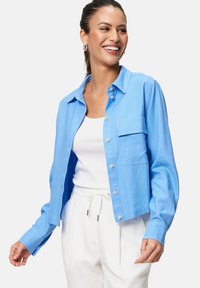 Chemise bleu clair à manches longues, avec une poche poitrine et une coupe courte, fabriquée dans un tissu lisse. Associée à un pantalon blanc.