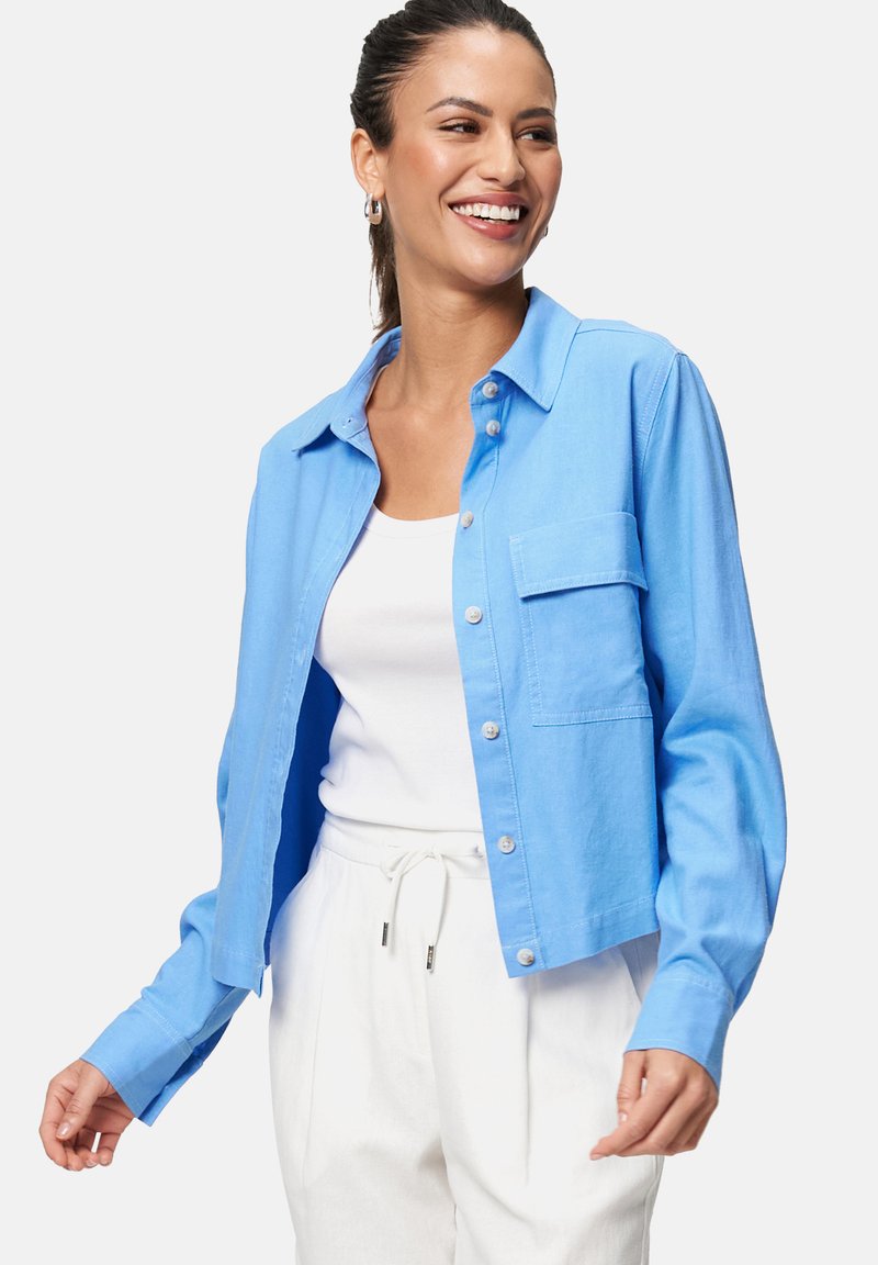 Chemise bleu clair à manches longues, avec une poche poitrine et une coupe courte, fabriquée dans un tissu lisse. Associée à un pantalon blanc.
