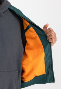 Kapjack met een donkergrijze buitenlaag, een contrasterende feloranje voering en groene ritsaccenten, die zachte stoftexturen onthullen.