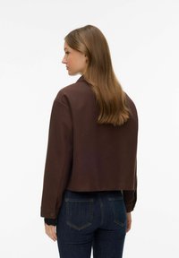 Femme aux longs cheveux bruns portant une veste marron courte et un jean bleu foncé, face à gauche sur un fond blanc uni.