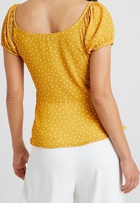 Blusa gialla senape con maniche a sbuffo e pois bianchi, dotata di scollatura quadrata e vita aderente. Tessuto testurizzato.