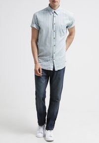 Hellblaues Kurzarmhemd mit weißen vertikalen Streifen, Knopfleiste, linke Brusttasche, kombiniert mit dunkelblauer Jeanshose und weißen Turnschuhen.