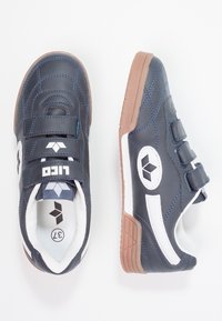 Baskets de sport navy avec des accents blancs, des sangles Velcro et une semelle en caoutchouc texturée. Présente un design matelassé et un détail de logo sur le côté.
