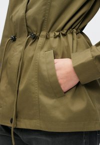 Πράσινο ελαφρύ μπουφάν με σφηκοφωλιά στη μέση, διαθέτει πλαϊνές τσέπες και λεπτομέρειες με κορδόνια, κατασκευασμένο από λείο ύφασμα με ματ φινίρισμα.