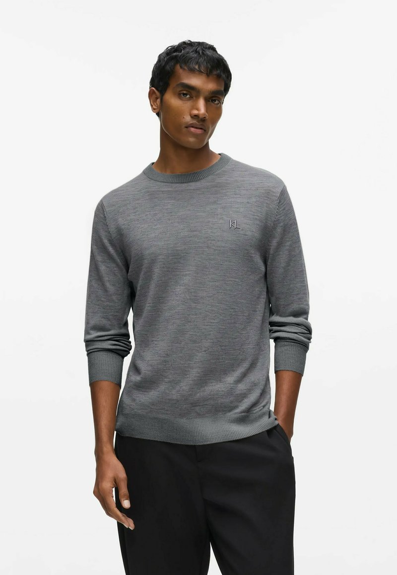 Homme portant un pull gris uni à col rond avec poignets et bas côtelés, associé à un pantalon noir, debout devant un fond blanc.