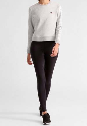 Femme portant un sweat-shirt à manches longues gris clair et des leggings noirs, marchant vers l'avant sur un fond blanc uni.