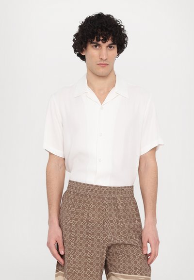 Camicia bianca a maniche corte con colletto, caratterizzata da una trama liscia. Abbinata a pantaloni corti marroni con un motivo geometrico e una cintura elastica.