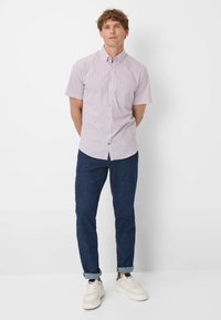 Camicetta a maniche corte con bottoni di colore viola chiaro, decorata con un piccolo motivo geometrico, abbinata a pantaloni blu scuro e sneakers bianche.