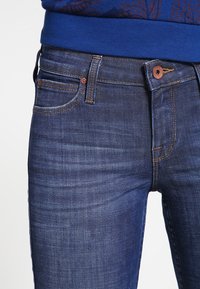 Primer plano de unos pantalones de mezclilla azul con bolsillos frontales y remaches de cobre, llevados con una blusa azul de estampado.