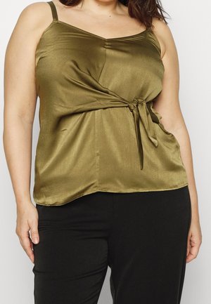 Femme portant un haut sans manches en satin vert olive avec un détail noué sur le côté, associé à un pantalon noir, visible du cou aux hanches.