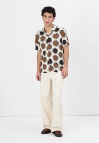 Camicia a maniche corte color crema con grandi soli decorati in marrone e nero. Indossata con pantaloni palazzo bianchi e scarpe scure.