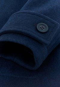 Dettaglio di cappotto in lana blu navy con tessuto strutturato, colletto arrotondato e bottone nero a due fori sul polsino.