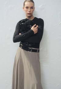 Top negro de manga larga, falda beige de cintura alta con pliegues y bolso de cuero negro. El conjunto incluye un cinturón negro con hebilla plateada.