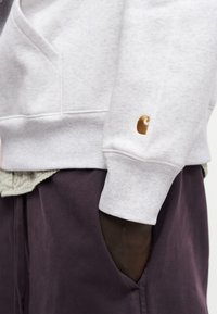 Χέρι με σκούρο χρώμα δέρματος στην τσέπη σκούρων μωβ παντελονιών, φοράει ανοιχτό γκρι φούτερ με μικρό καφέ λογότυπο στη μανσέτα του μανικιού.