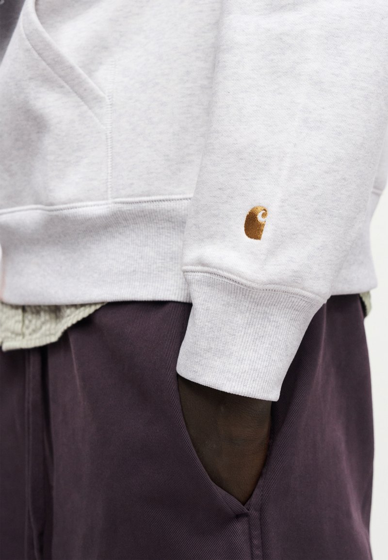 Donkere hand in de zak van donkerpaarse broek, met een lichtgrijze trui met een klein bruin logo op de mouwmanchet.