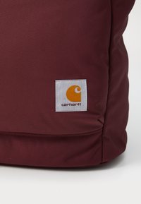 Bordeaux ryggsekk med teksturert stoff, som har en firkantet hvit lap med en oransje-og-gul logo i nedre hjørne.