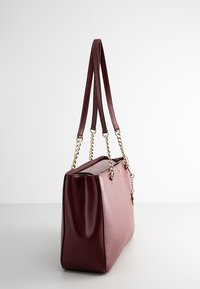 Sac à main en cuir rouge foncé avec chaîne dorée et bandoulières en cuir, posé debout sur une surface blanche unie.