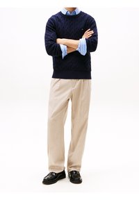 Pull en maille torsadée bleu marine porté sur une chemise rayée bleu clair, pantalon beige et mocassins noirs ; présente des poignets contrastants et un petit détail logo.