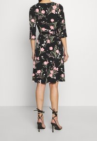 Blumenbedrucktes Kleid mit Dreiviertelärmeln, schwarzem Hintergrund und rosa Blumen. Hat eine taillierte Taille und einen ausgestellten Rock. Trägt schwarze Riemchen-Absätze.