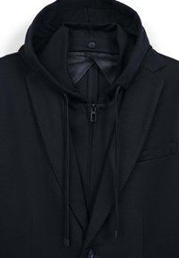 Schwarzer Blazer über einem schwarzen ZIP-Hoodie mit Kapuze und Kordelzügen, von der Brust aufwärts vor einem weißen Hintergrund gezeigt.