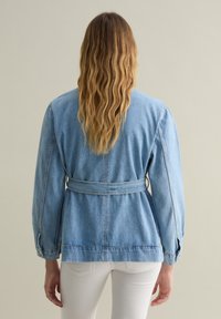 Giacca in denim di un blu chiaro con vita regolabile, maniche lunghe e vestibilità comoda. Caratterizzata da dettagli di cucitura e polsini delle maniche elasticizzati.