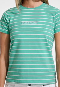 Žalias dryžuotas marškinėlis su šviesiai rožiniais horizontaliais dryžiais, pagamintas iš minkštos medžiagos. Priekyje turi baltą "GUESS JEANS" logotipą.