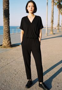 Mlada žena u crnom kombinezonu i tenisicama stoji na šetnici uz more s palmama pod vedrim nebom.