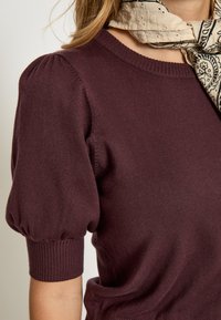 Vínově červený svetr s krátkým rukávem, kulatým výstřihem, texturovaným pletením a nabíranými rukávy. Má žebrované manžety na koncích rukávů.