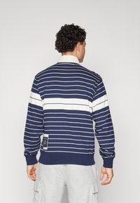 Navy striped sweatshirt with white horizontal stripes, long sleeves, and a collar. Features a logo patch on the lower side. Soft fabric texture.