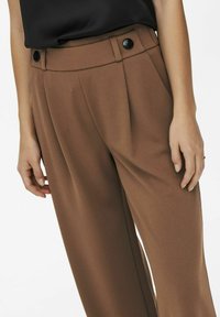 Personne portant un pantalon marron taille haute avec des boutons noirs et un haut noir rentré, les mains détendues le long du corps.
