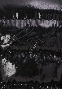 Crni paillet tkanina s različitim teksturama, koja sadrži izvezeni logo crnom niti. Površina reflektira svjetlost i stvara shimmer efekt.