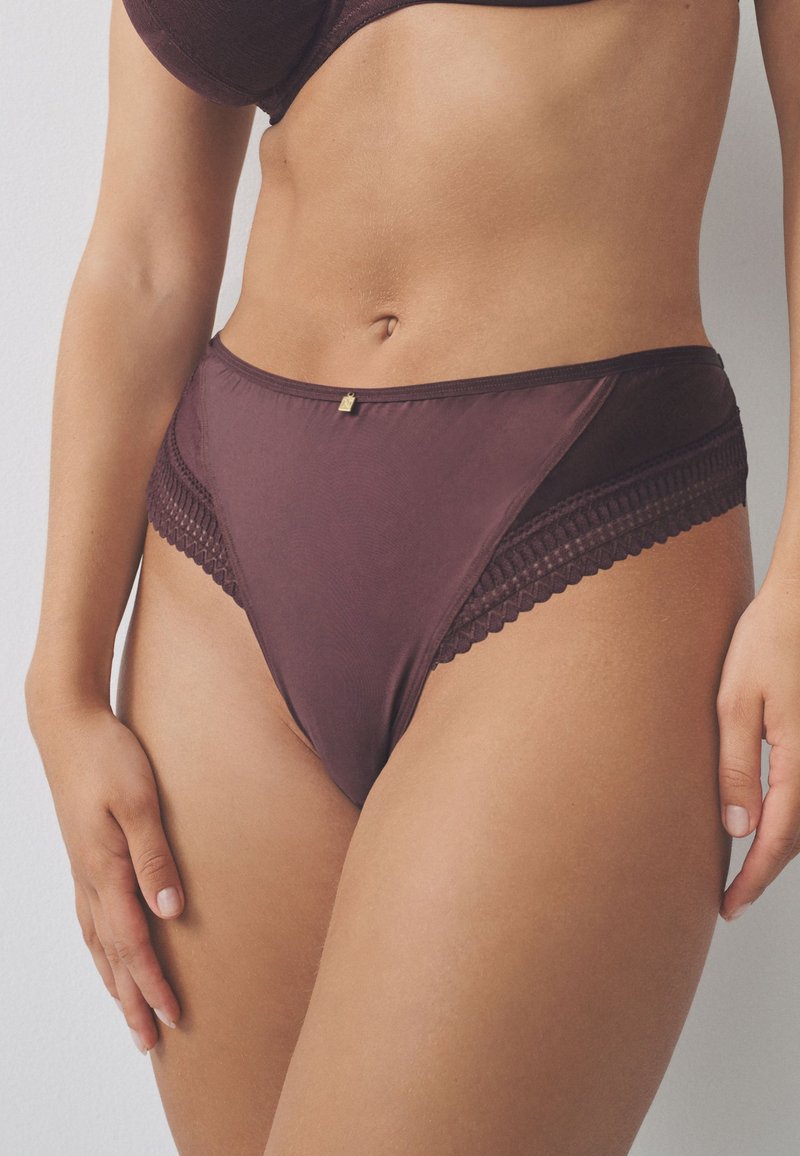 Culotte taille haute marron foncé avec une texture lisse et des accents en dentelle sur les côtés. Présente un petit emblème doré à l'avant.