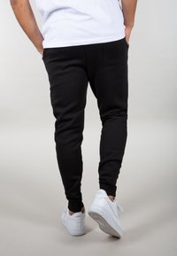 Person med sorte joggers og hvide Nike Air sneakers står med hænderne i lommerne på en lys baggrund.