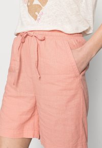Shorts en lin rose clair avec une taille élastique, cordon de serrage ajustable et poches latérales. Texture lisse, design décontracté.