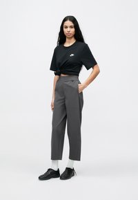 Fekete, rövidített Nike póló elöl csomózva, szürke, szabott nadrággal és fekete cipővel párosítva. A modell egyik kezét a zsebébe helyezi.