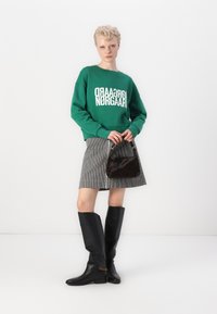 Felpa verde con testo bianco, gonna a righe, stivali neri fino al ginocchio e piccola borsa marrone. Materiali lisci e chiari elementi di design.