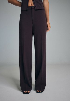 Personne portant un pantalon marron foncé taille haute à jambes larges avec une fermeture à boutons, accompagné de sandales noires à bouts ouverts sur un fond uni.