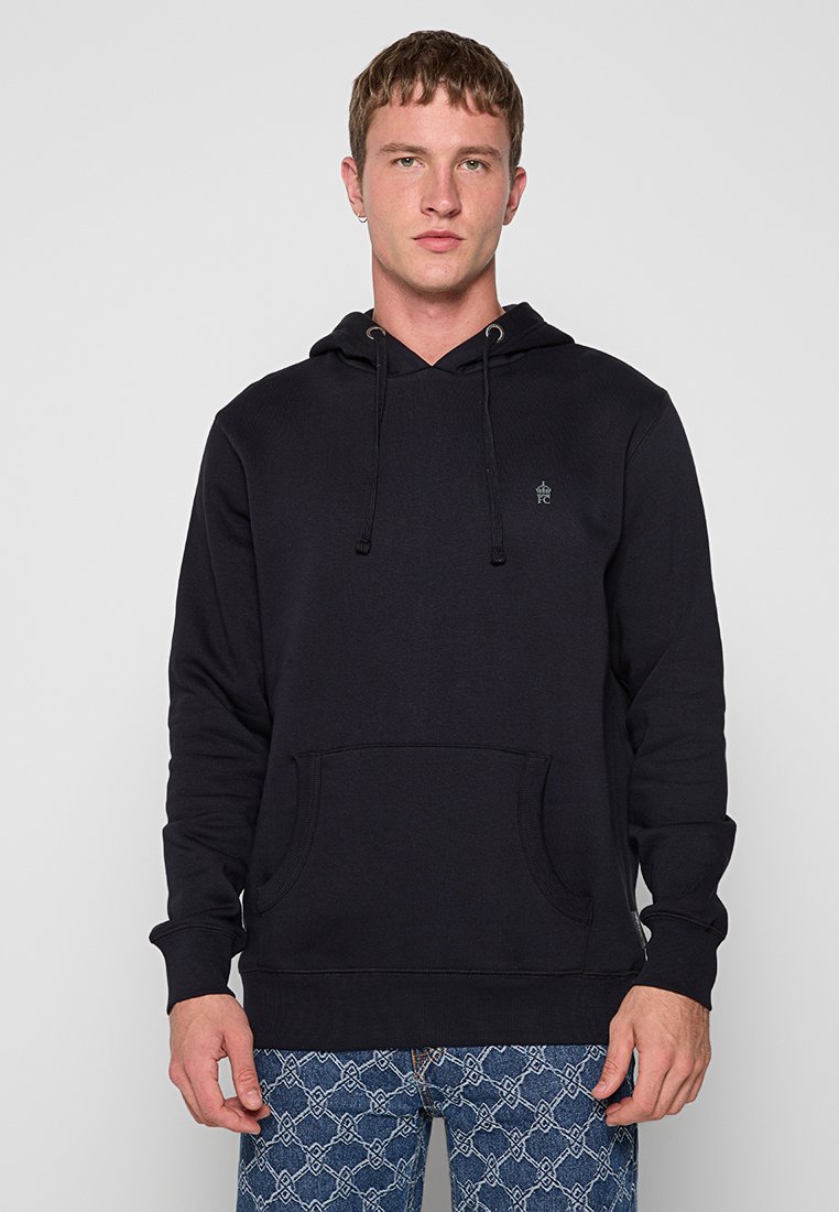 Junger Mann in einem schwarzen Hoodie mit Vordertasche und gemusterten blauen Jeans vor einem schlichten, hellen Hintergrund.
