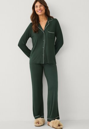 Kvinde smiler, iført mørkegrøn pyjamas med knapper og hvide kanter samt beige fluffy hjemmesko, står foran en ensfarvet lys væg.