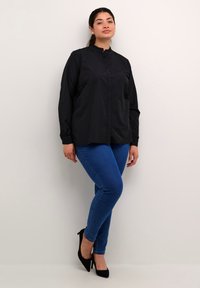 Sort langærmet bluse med knaplukning foran og høj krave, parret med blå skinny jeans og sorte højhælede sko, stående mod en hvid væg.